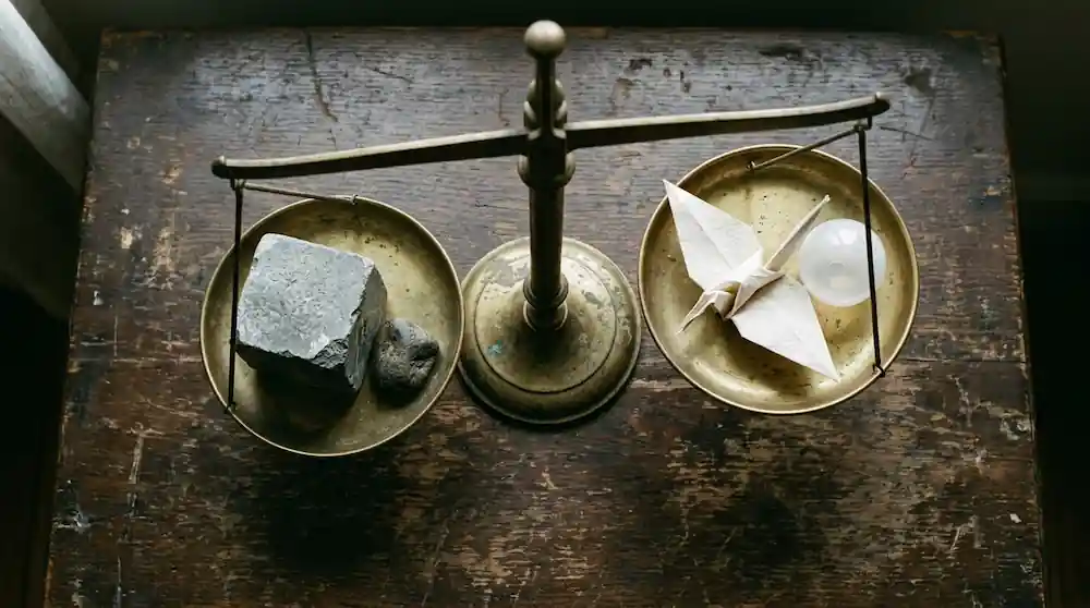 Overhead flat-lay of four differently weighted objects on an old brass balance scale - a solid stone cube, a smaller stone, a hollow plastic shell, and a paper origami shape - uneven lighting, documentary minimalism, muted palette, implying different amounts of substance.